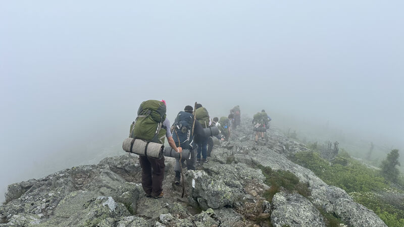 The image shows a group of hikers walking along a rocky ridge in a foggy environment. They are carrying backpacks and appear to be on a challenging hike. The fog obscures the background, creating a sense of mystery and adventure. The hikers are focused on their path, navigating the uneven terrain.